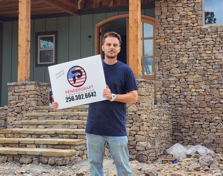 Beau Pendergraft holding a Pendergraft Construction sign in front of a completed stone and timber home in Guntersville, Alabama