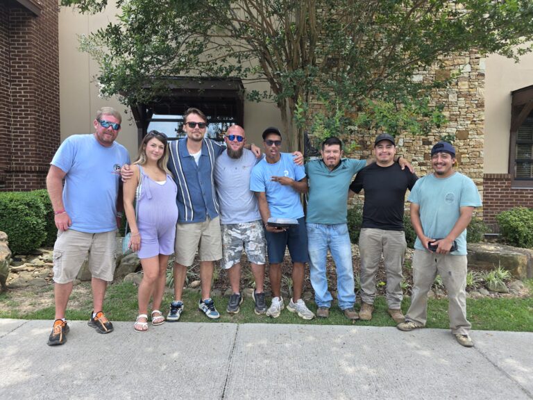 Group of eight people standing arm-in-arm on a sidewalk in front of a building with brick and stone accents, smiling for a team photo under a tree, several wearing matching Pendergraft-branded shirts.